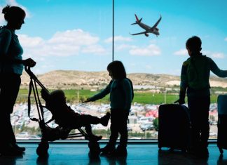 The silhouette of a mom with her toddler child and two older kids at an airport, standing with luggage.