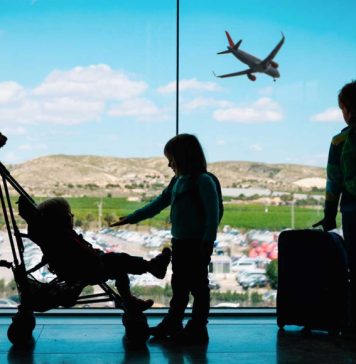 The silhouette of a mom with her toddler child and two older kids at an airport, standing with luggage.