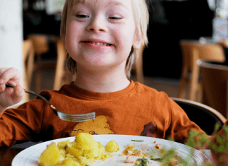 A little boy with Dow Syndrome in an orange shirt uses a fork to eat a meal while sitting in a chair at the table.
