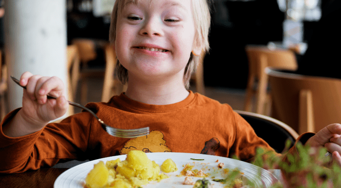 A little boy with Dow Syndrome in an orange shirt uses a fork to eat a meal while sitting in a chair at the table.