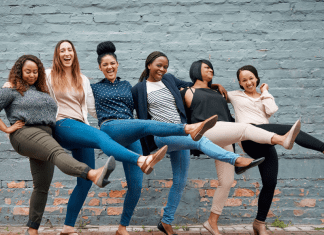 Six women of all types of skin colors locking arms and kicking one leg in the air, laughing