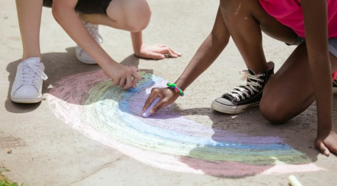 Two kids draw a rainbow with sidewalk chalk.