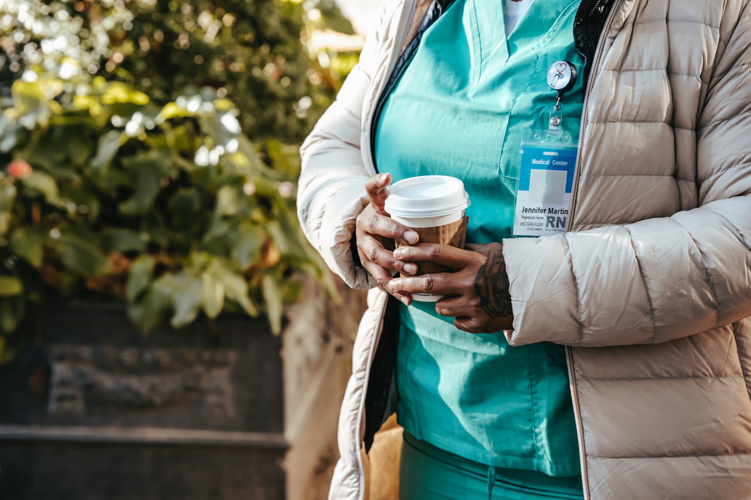 A nurse in scrubs holds a cup of coffee while taking a walk outside.