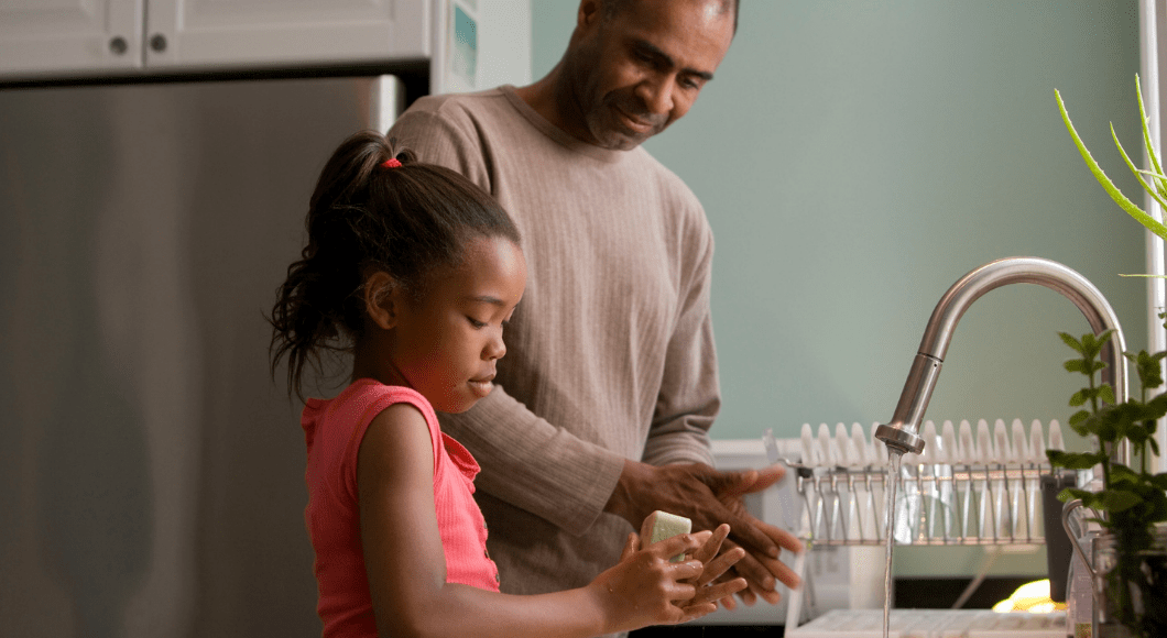 A dad helps his daughter wash her hands at the kitchen sink.
