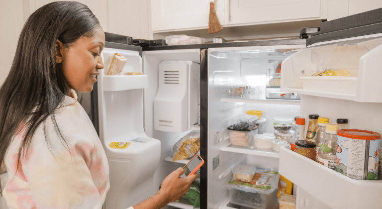 A woman cleans out her fridge and looks at her phone.