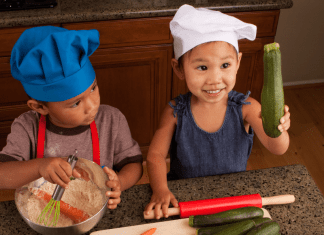 Two toddler cooking in the kitchen. One holds up a cucumber.