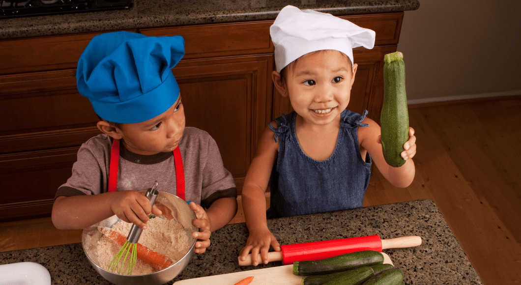 Two toddler cooking in the kitchen. One holds up a cucumber.