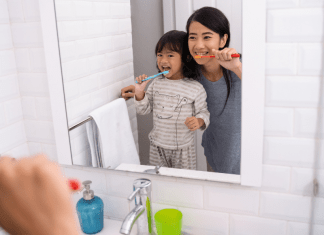 A mom and daughter brush their teeth in the bathroom mirror.