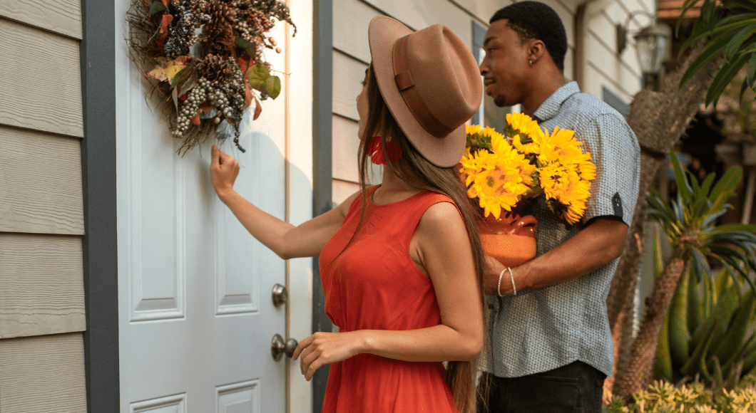 A couple, the man holding a pot of sunflowers, arrives at the front door with a wreath on it as the woman begins to knock. 