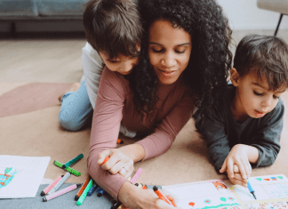 A mom and her kids draw in a coloring book together on the living room floor.