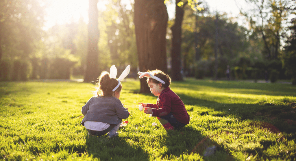 Two children wearing bunny ears hunt for Easter eggs in the park.