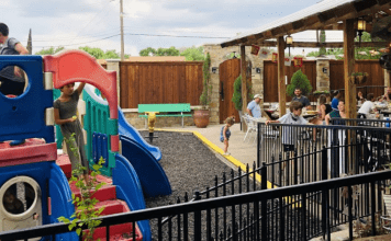 Kids play on a playground adjacent to a restaurant's outdoor patio where the grownups sit, eat, and watch the kids.